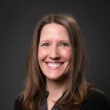 Smiling woman with long brown hair, wearing a dark floral shirt and cardigan.