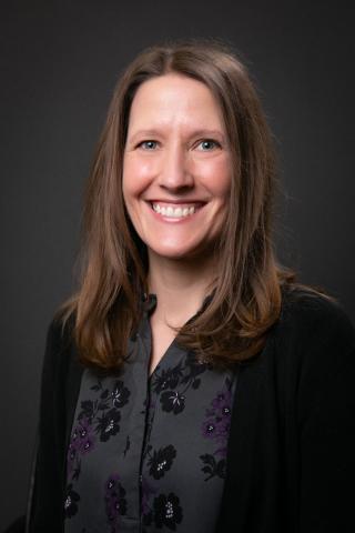 Smiling woman with long brown hair, wearing a dark floral shirt and cardigan.