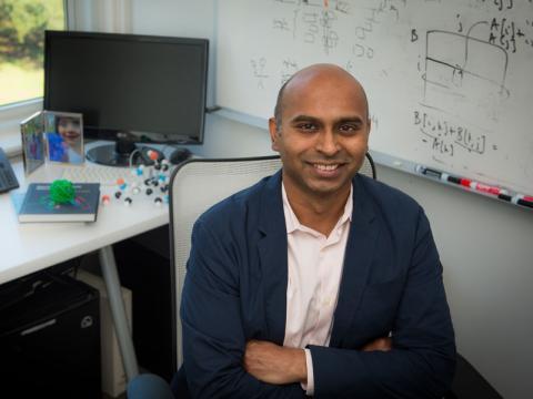 Smiling man with crossed arms in an office, scientific models and equations behind him.